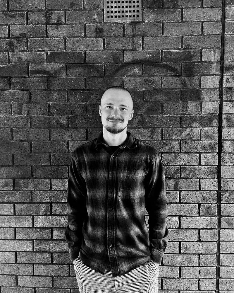 A smiling man standing in front of a textured brick wall, wearing a dark plaid shirt and checkered pants.